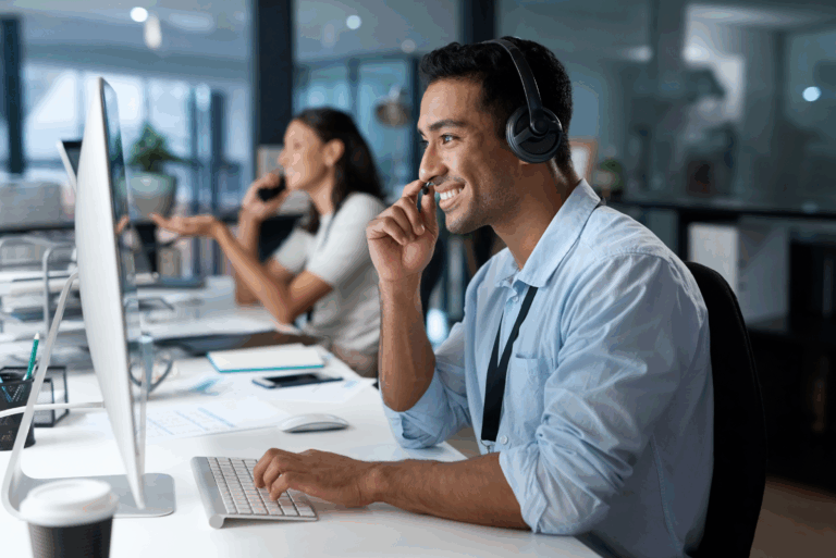 A man wearing a headset is smiling while talking on the phone and working on a computer at a desk in a modern office setting.